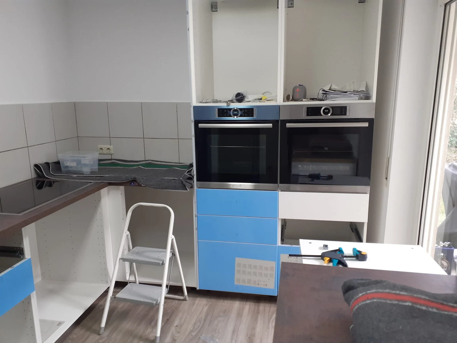 Weiße Küche während der Montage mit zwei Einbaugeräten, blauer Schutzfolie und Trittleiter
